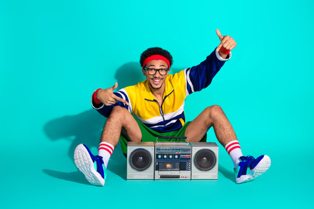 Energetic young man wearing retro sportswear sitting on teal background holding a vintage boombox, showcasing active lifestyleの写真素材