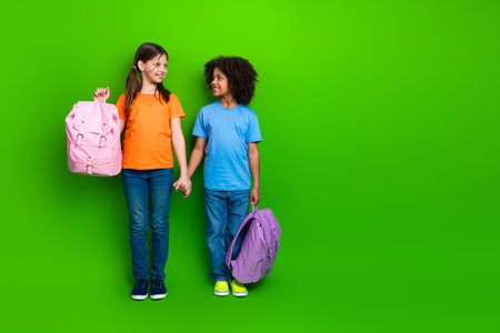 Smiling schoolgirls with colorful backpacks holding hands on a vibrant green background, symbolizing childhood friendship and diversityの写真素材