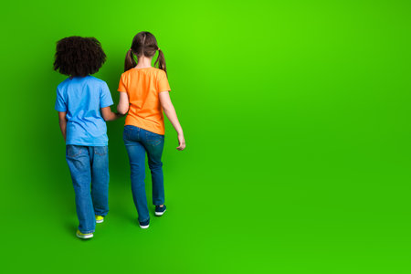 Two young girls holding hands, walking together against a vibrant green background, symbolizing friendship and diversityの写真素材