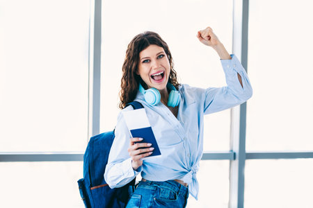 Excited young woman celebrating with a passport and headphones at the airport, ready for her travel...