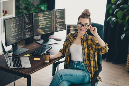 Young female programmer working remotely in a modern home office with multiple monitors and a...