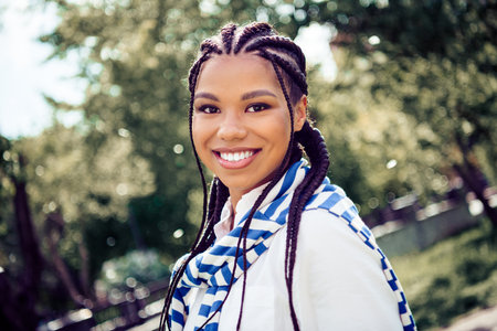 Young woman with braided hair enjoying a sunny day outdoors in a park, showcasing her cheerful smile and fashionable attireの写真素材
