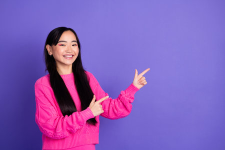 Cheerful young woman in a vibrant pink sweater points excitedly against a bold purple background, exuding positivityの写真素材