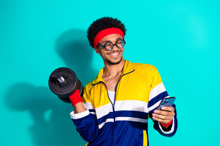 Young afro haired man wearing retro sportswear smiling with weight in hand and using smartphone