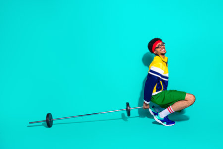 Young man in retro sportswear squatting with barbell against teal background showcasing vintage gym training and funky activewear styleの写真素材