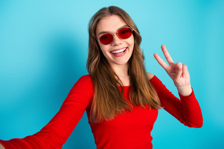 Cheerful Young Woman in Red Top and Sunglasses Posing Against Blue Backdrop, Showing Victory Sign