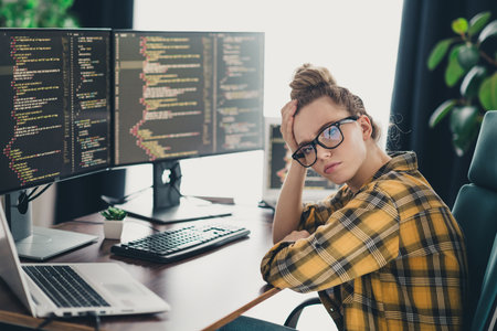 Young female programmer working from home on coding tasks, wearing casual plaid shirt and glasses, looks thoughtfulの写真素材