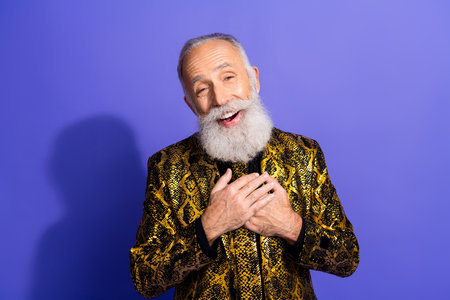 Cheerful mature man in golden jacket with hands on chest expressing delight against a vibrant purple backgroundの写真素材