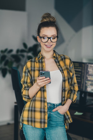 Qualified professional woman with glasses using smartphone in creative workspace, wearing casual attire, smiling confidentlyの写真素材
