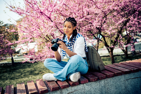 Charming woman in a park enjoying a spring day with a camera under blooming pink trees, appreciating natureの写真素材