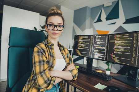 Young female programmer working in a casual environment with a dual-monitor desktop setup, featuring...