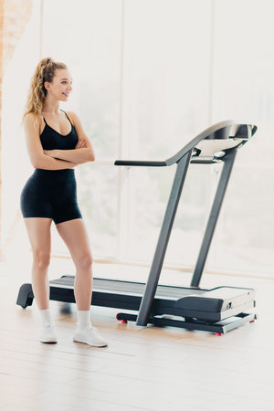 Active woman in activewear standing confidently near treadmill during a home workout session in a...