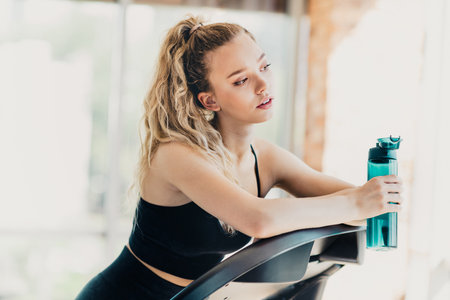 Young athletic woman resting on treadmill holding water bottle inside a bright gym interior...