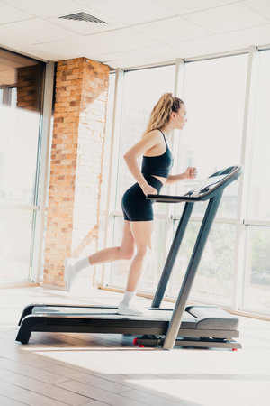Young woman exercising on a treadmill in a modern gym setting with bright natural lighting,...