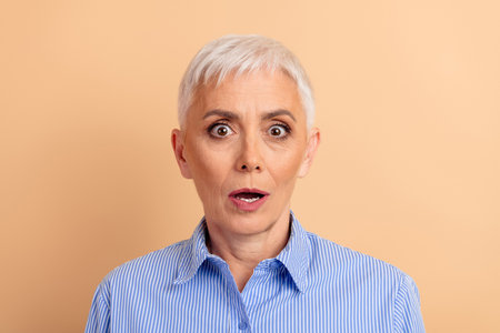 Surprised mature woman with short white hair wearing a blue striped shirt against a beige background showcasing a shocked expression.の写真素材
