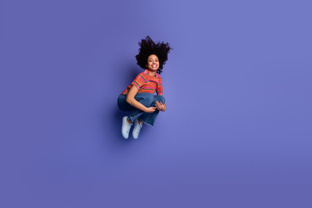 Joyful young woman jumping on purple background wearing casual outfit embracing vibrant and stylish...