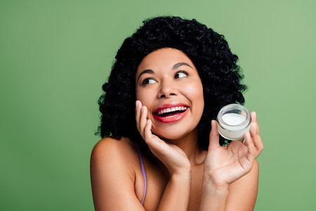 Beautiful young woman joyfully posing with a skincare cream jar against a green background, promoting self-careの写真素材