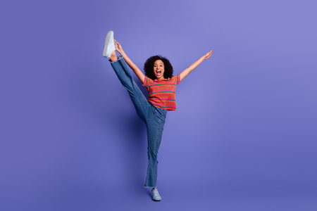 Energetic young woman in casual attire enthusiastically posing with raised leg on a vibrant purple backgroundの写真素材