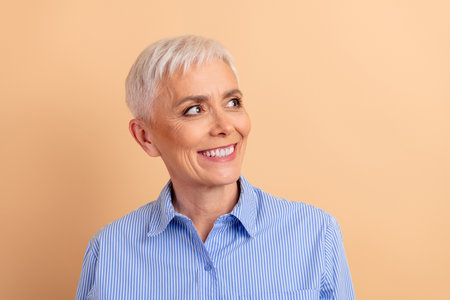 Smiling mature woman with short white hair and blue striped shirt on beige background, expressing...