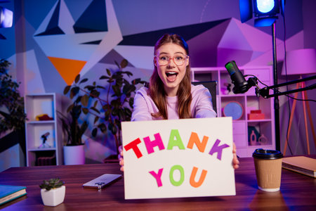 Young female blogger holding a thank you sign during a colorful recording session with studio lights...