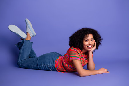 Young woman lying casually on purple background in stylish outfit, smiling warmly, expressing...