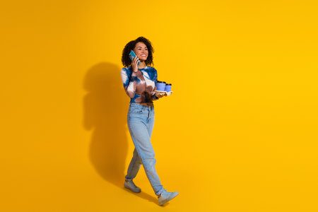 Young woman walking confidently, talking on the phone and holding a gift on a vibrant yellow...