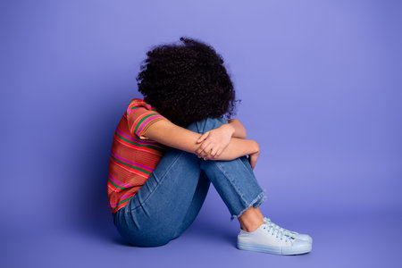 Young woman in casual outfit sitting against purple background, embracing her legs and portraying...