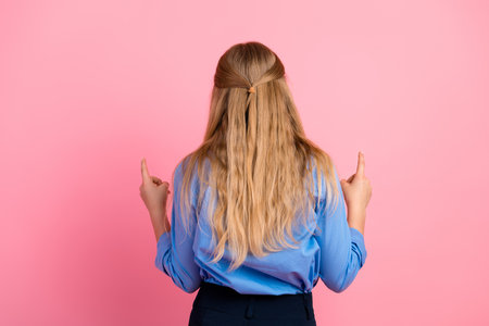 Young girl with blonde hair gesturing thoughtfully, standing against a pink background wearing stylish blue shirtの写真素材