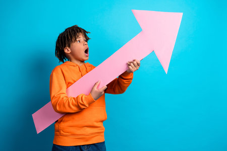 Delighted boy in orange hoodie holds a pink arrow prop, showcasing enthusiasm against a bright blue backgroundの写真素材