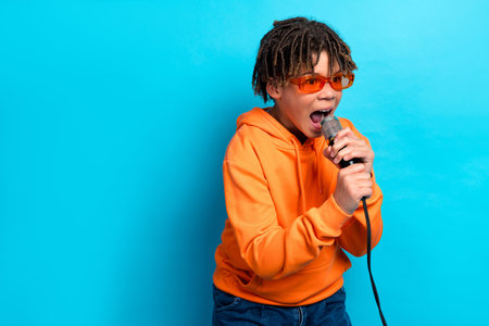 Energetic young boy karaoke singing with microphone in orange hoodie against vibrant blue backgroundの写真素材
