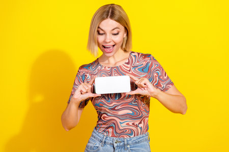 Cheerful young woman showing smartphone with excitement in colorful top against vibrant yellow backgroundの写真素材