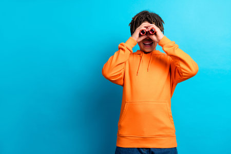 Cheerful boy in orange hoodie playfully looking through improvised binoculars against vibrant blue backgroundの写真素材