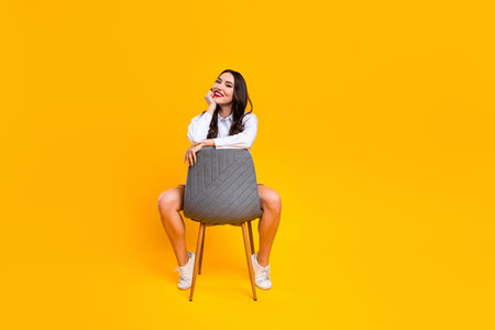 Charming woman sitting stylishly on a chair against a yellow background representing leisure, lifestyle and modern fashionの写真素材