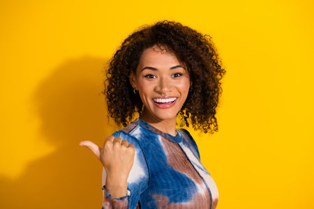 Young woman with curly hair wearing a colorful print top in front of a bright yellow background with a cheerful smileの写真素材