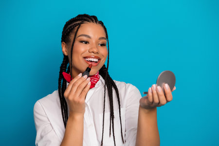 Smiling young woman applying lipstick while holding a compact mirror, standing against a vibrant blue backgroundの写真素材