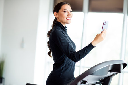 Athletic woman in activewear using a smartphone while training on a treadmill in a modern, bright...