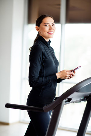 Woman in sporty outfit smiling while using her smartphone on a treadmill, enjoying a healthy...