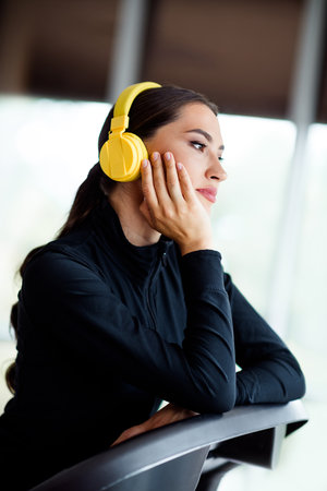 Active woman in black activewear indoors wearing yellow headphones and thinking by a window, highlighting sport and fitnessの写真素材