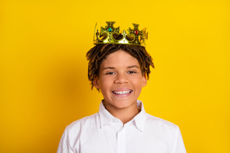 Smiling boy with gold crown stands against yellow background representing joy, confidence, and childhood happinessの写真素材