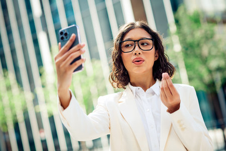 Confident young businesswoman taking a selfie outdoors while discussing opportunities, dressed in formalwear in a modern urban setting.の写真素材