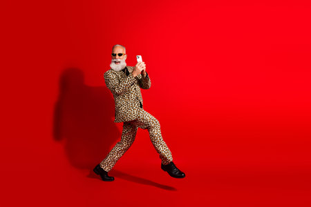 Stylish elderly man in leopard print suit holding a phone on a red background exuding charm and modern charismaの写真素材