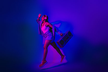 Vibrant young woman dancing with boombox under neon lights wearing a casual denim outfit exemplifies...