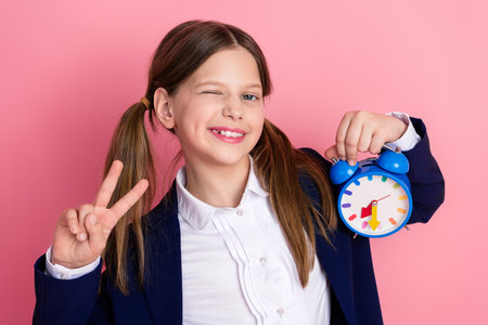 Smart young schoolgirl holding colorful alarm clock and showing peace symbol on pink background