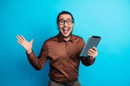 Enthusiastic young man holding digital device against teal backdrop, expressing excitement and joy with positive energyの写真素材