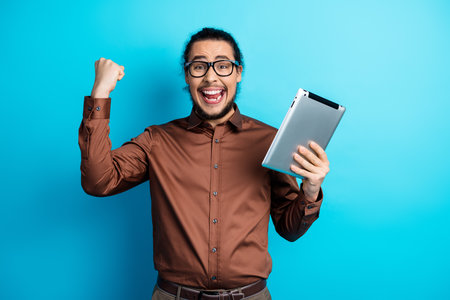 Excited young professional man enjoying success with a tablet against a vibrant blue background,...