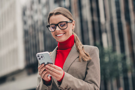 Stylish professional in elegant attire checking her phone outdoors in urban city against modern office buildingの写真素材