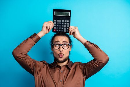Young professional man in brown shirt holding calculator on head with playful expression on teal backgroundの写真素材