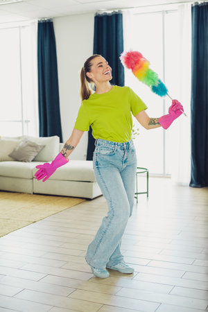 Young woman joyfully cleaning with colorful duster in modern living room, embracing positivity and...