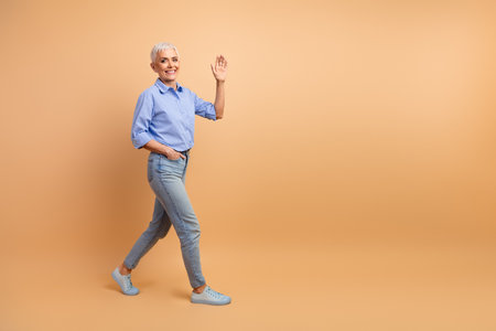 Confident mature woman in stylish casual attire gesturing with a friendly wave against a solid beige backgroundの写真素材