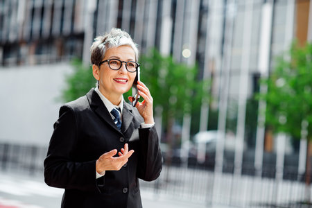 Confident mature businesswoman with short hair smiling while speaking on a mobile phone outdoors in...